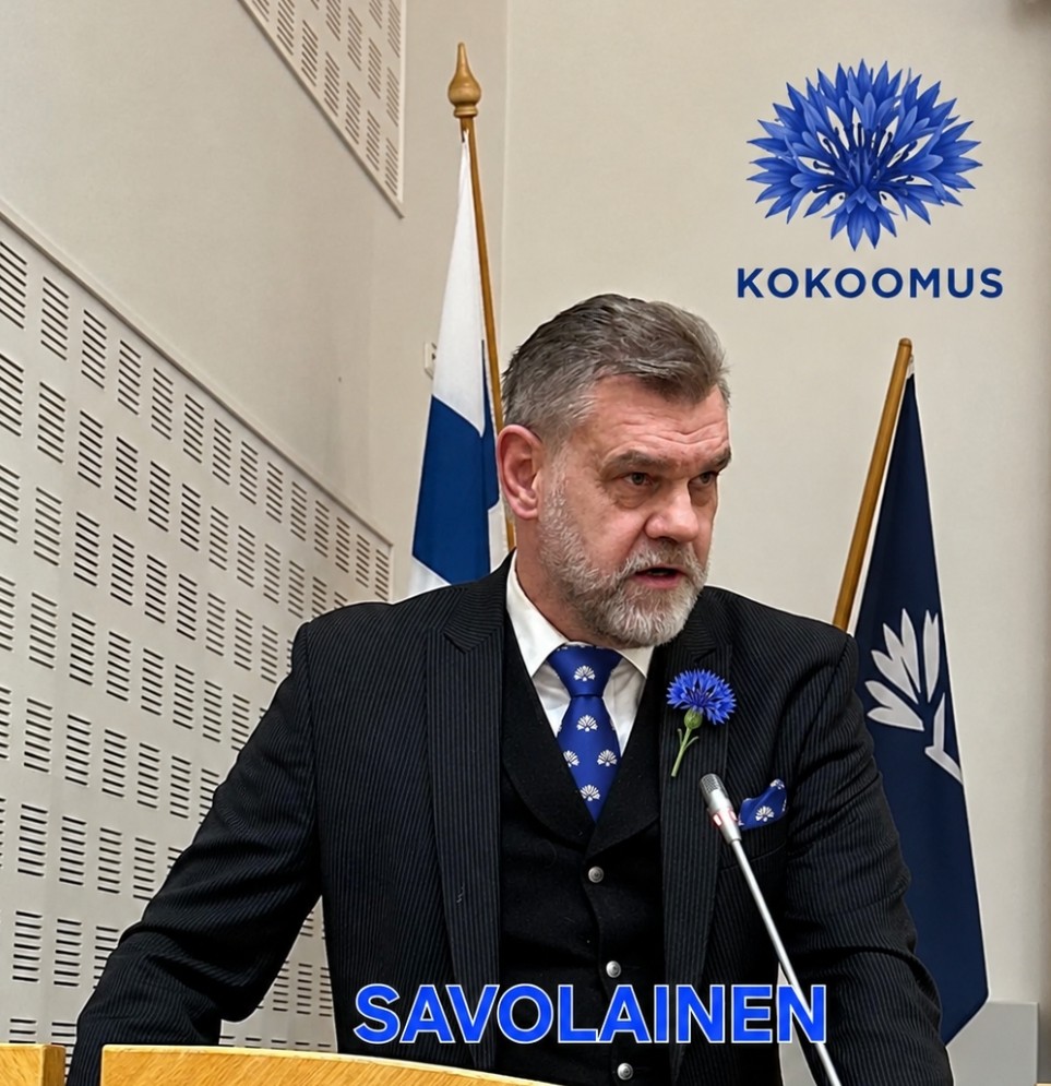 Jukka Savolainen puhujapöntössä, taustalla Suomen lippu ja Kokoomuksen tunnus